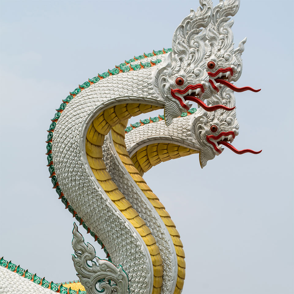 Ornate white serpent sculpture with three heads, red tongues, and golden scales on the underside against a cloudy sky