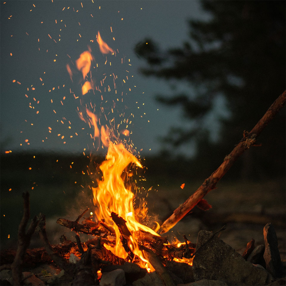 Close-up of a glowing campfire at dusk, with flames rising and sparks flying into the air. Surrounded by rocks and branches, with a dark forest and evening sky in the background.